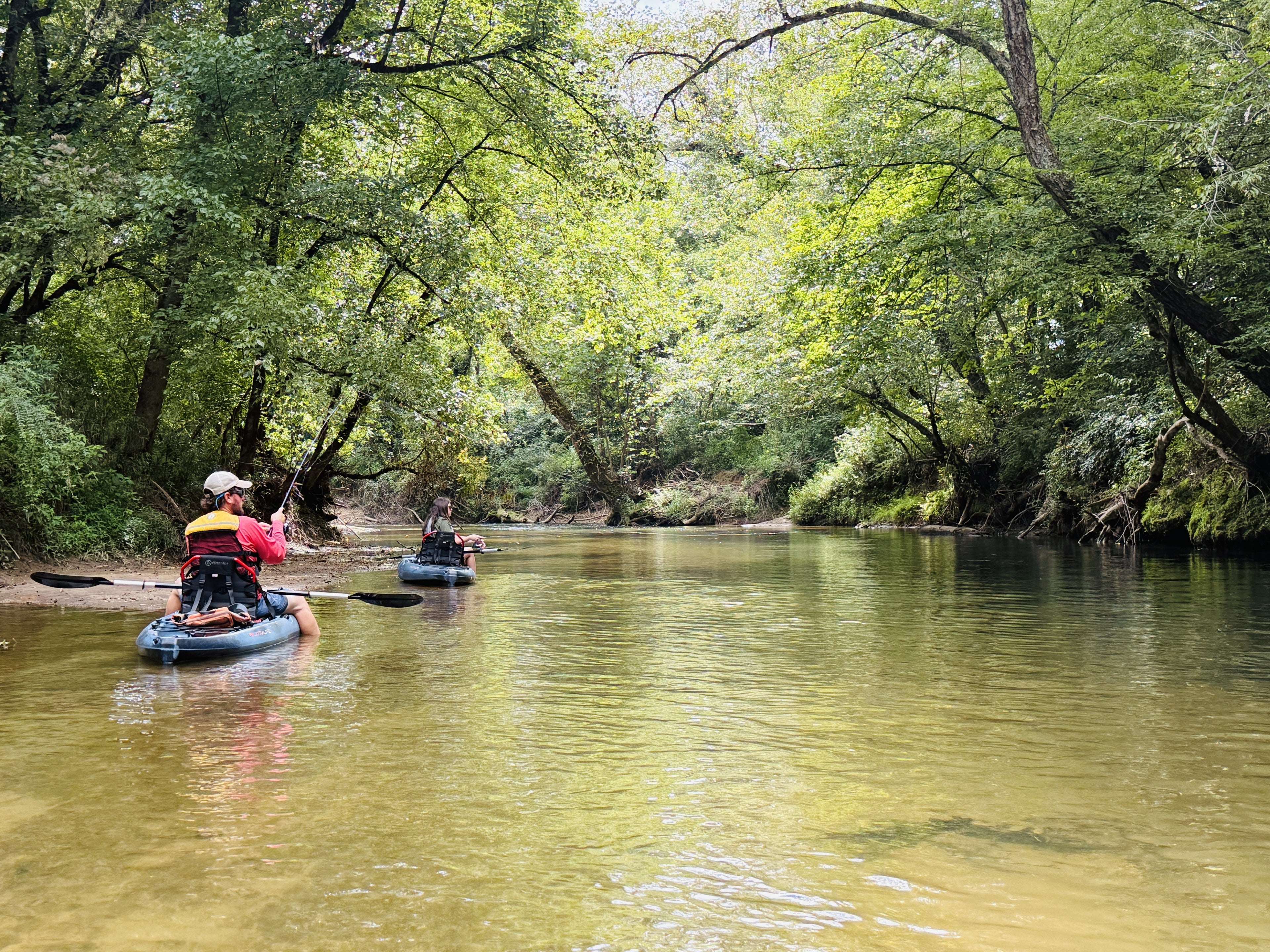 Two people kayaking and fishing on the Yadkin River surrounded by lush green trees.
